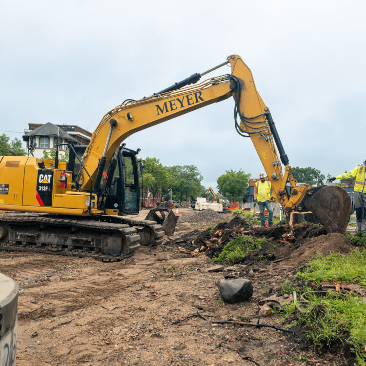 Yellow Meyer excavator clears debris; four workers supervise nearby.