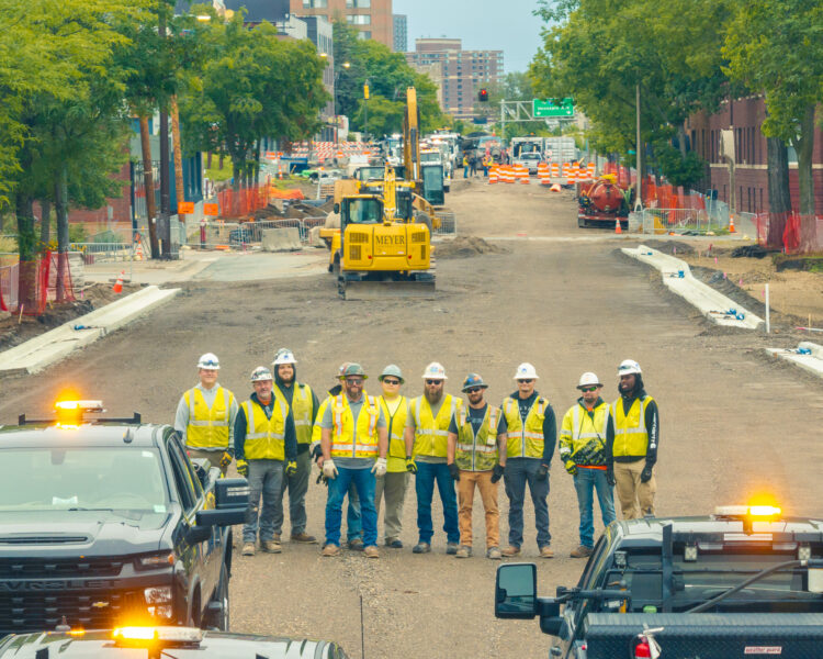 Construction workers in safety gear stand on a partially paved road.