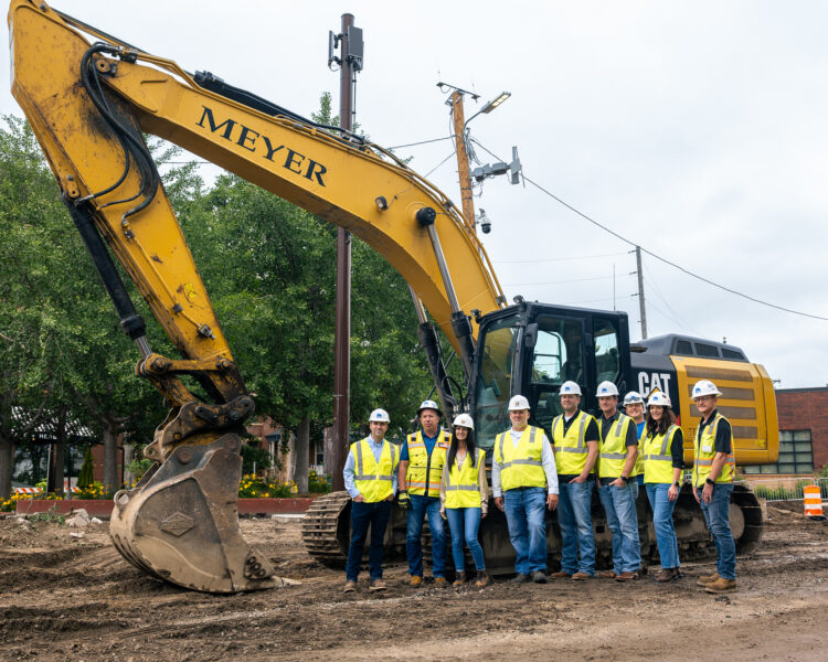 Workers in safety gear stand before a yellow excavator.