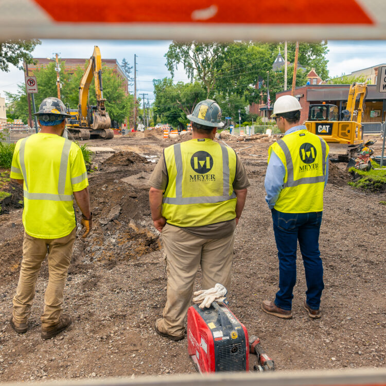 Three workers in safety gear watch roadwork with heavy machinery.