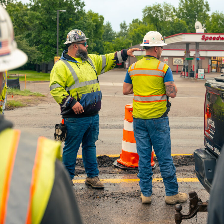 Construction workers gather near roadwork; Speedway gas station behind.