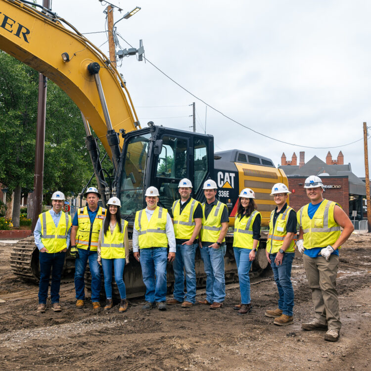 Nine construction workers pose by an excavator at a worksite.