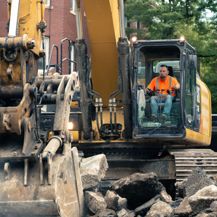Worker breaks concrete with yellow excavator near brick building.