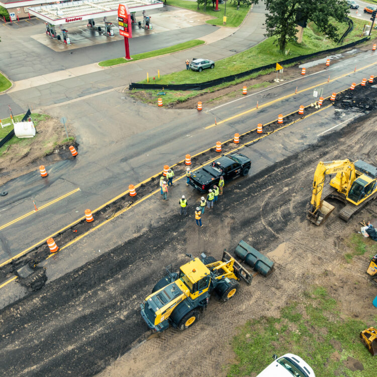 Aerial view: road construction, machinery, workers, cones by gas station.