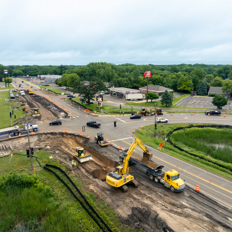 Aerial of roadwork, machines, traffic, buildings, and trees nearby.