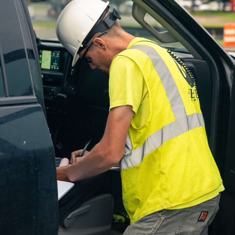 Construction worker notes on clipboard by open vehicle door.