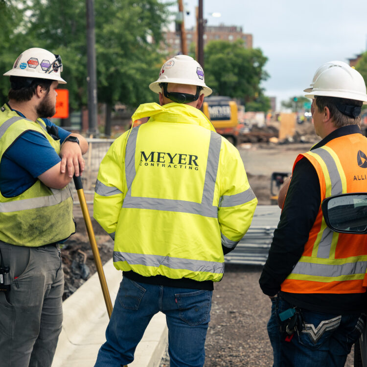Three construction workers in safety gear talk by roadwork.