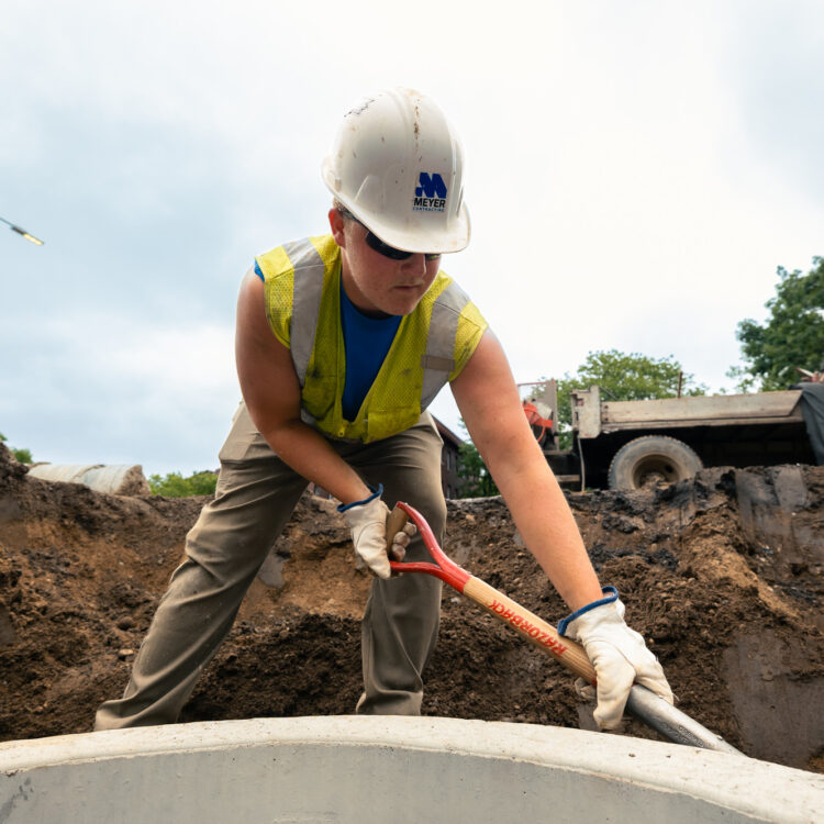 Worker in hard hat shovels mud at busy construction site.