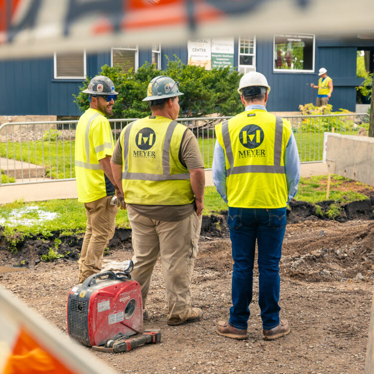 Three workers in vests stand near equipment; barriers and another worker.
