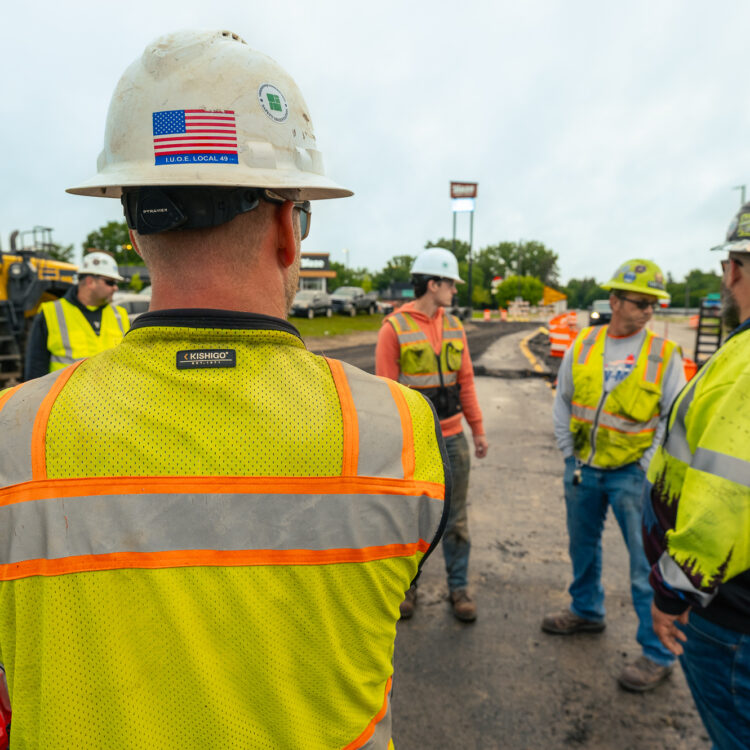 Construction workers in safety gear talk at site with equipment.