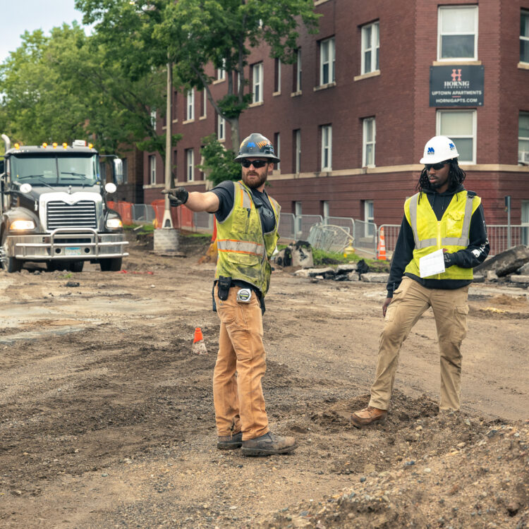 Two construction workers discuss plans on a city street.