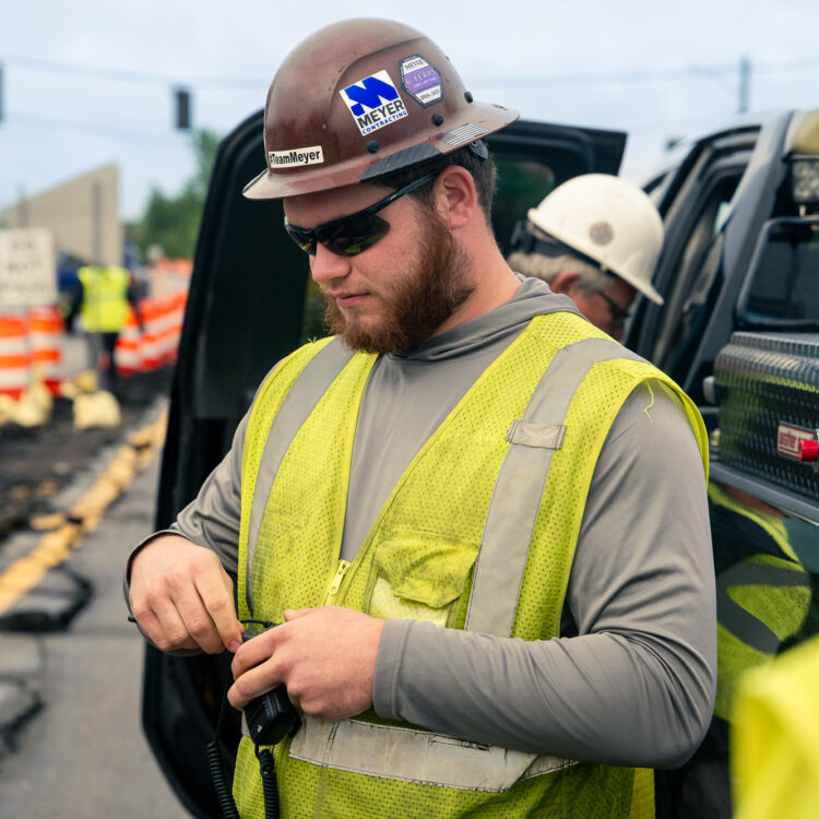 Worker in vest adjusts gear by truck at roadwork site.
