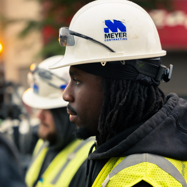 Worker in hard hat and vest stands outside; equipment behind.