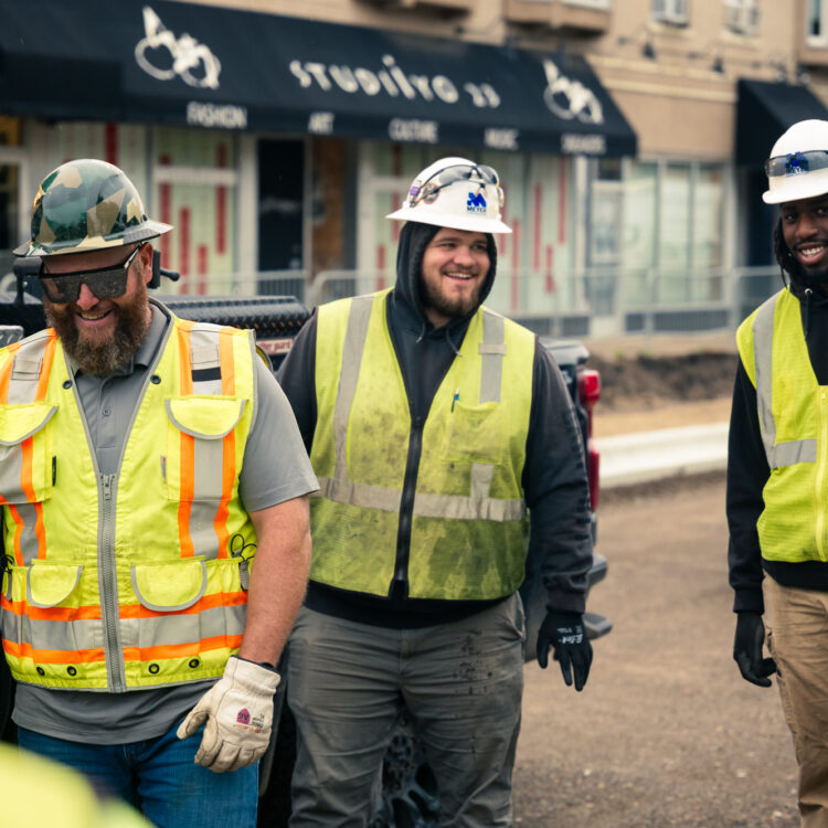 Three construction workers in safety gear smile near storefronts.