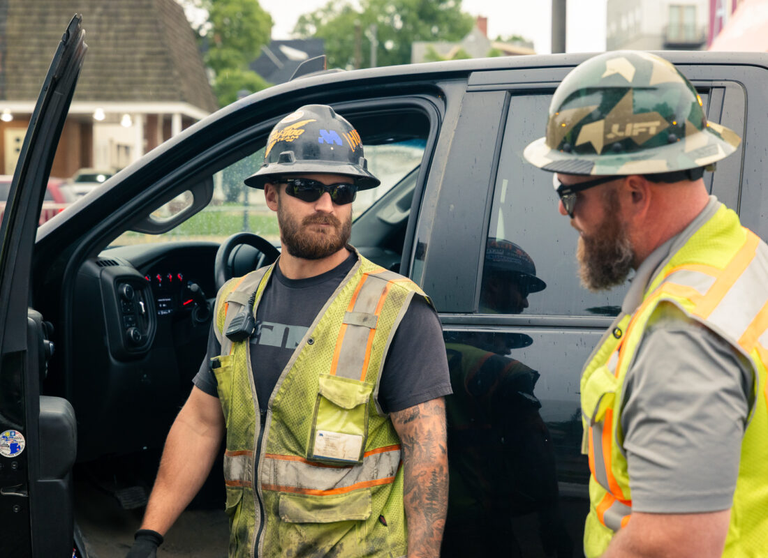 Two workers in safety gear talk by a black pickup.