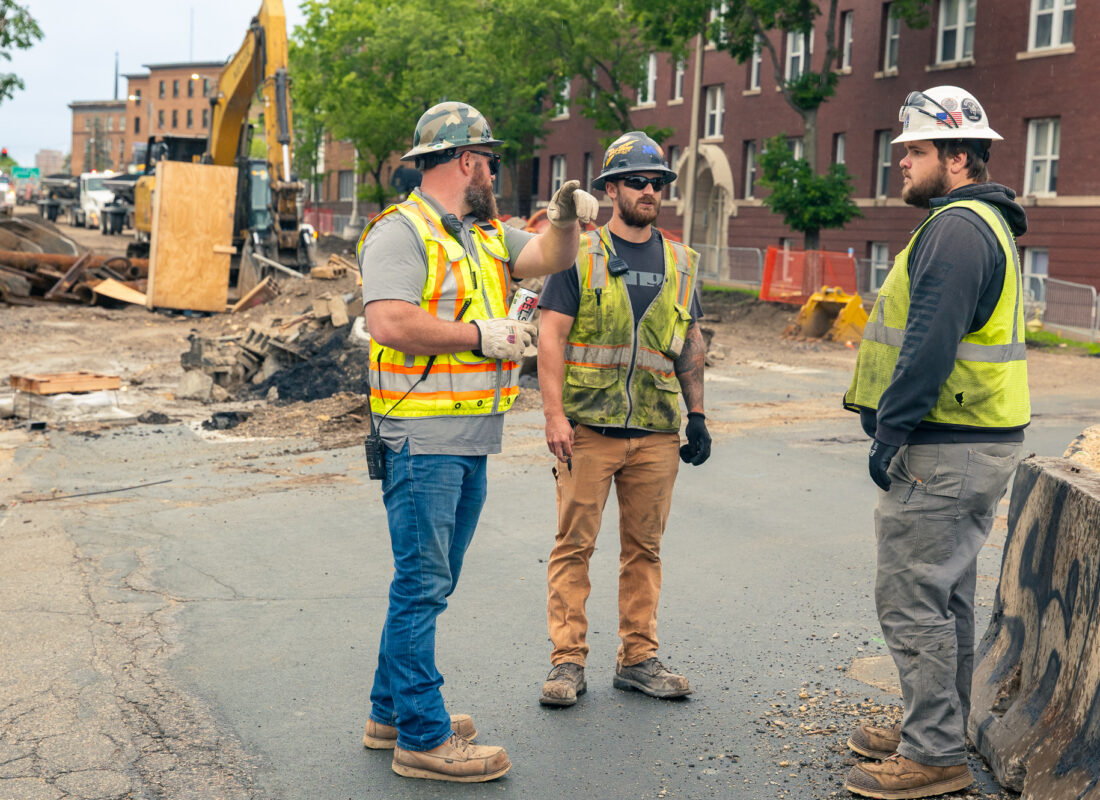 Three construction workers in safety gear talk by construction equipment.
