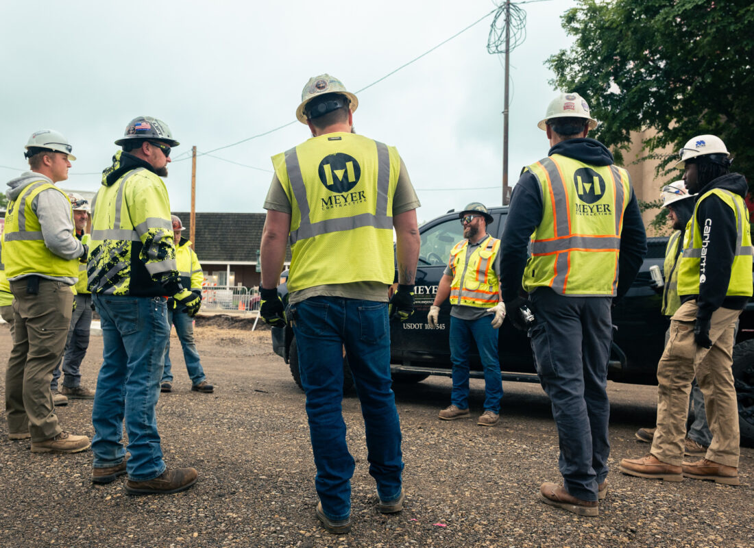 Construction workers in vests and helmets discuss outdoors in circle.