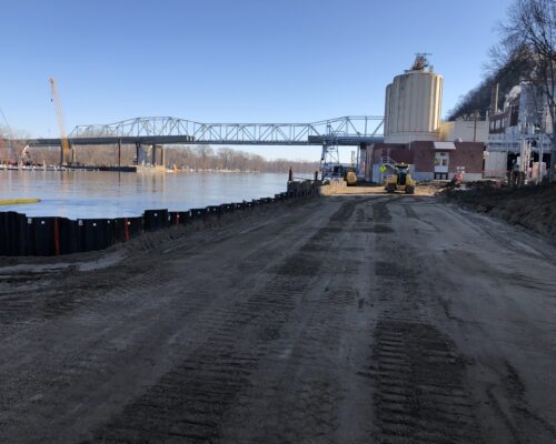 Riverbank construction: heavy machinery, sheet piling, bridge, clear day.