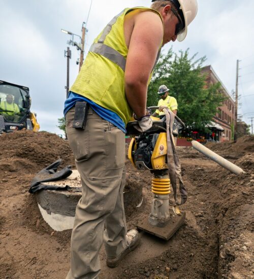 Worker operates compactor; another stands nearby on dirt site.