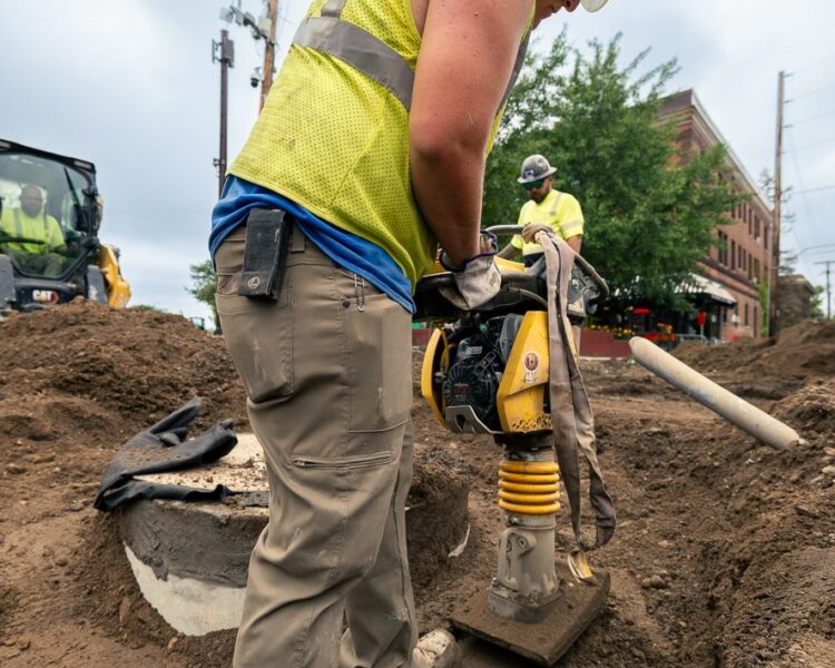 Worker operates compactor; another stands nearby on dirt site.