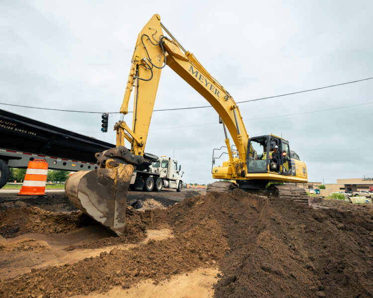 Yellow excavator digs near road; dump truck and barrels nearby.