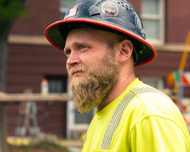 Bearded man in hard hat, neon shirt, stands by brick building.