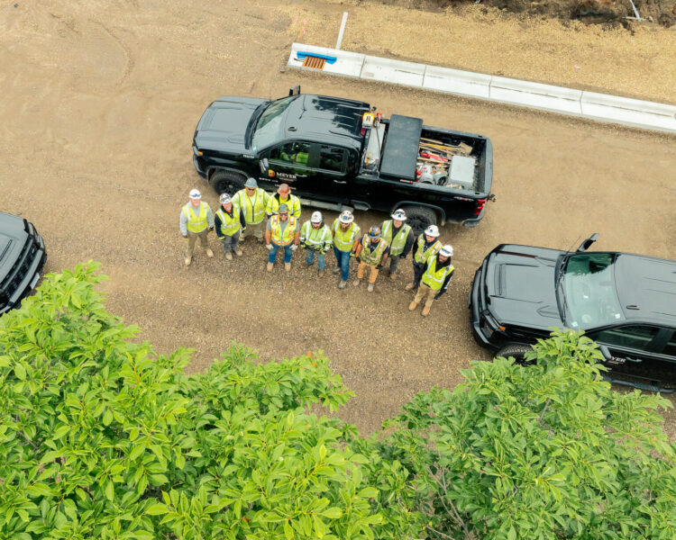 Construction workers in safety gear stand between trucks, viewed aerially.