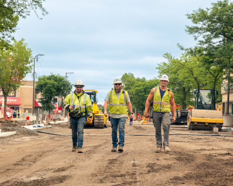 Three construction workers walk down an unpaved street under construction.