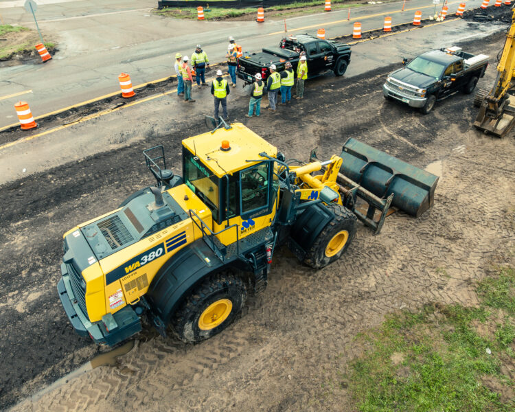 Aerial view: yellow loader, workers, trucks, roadwork, orange barrels.